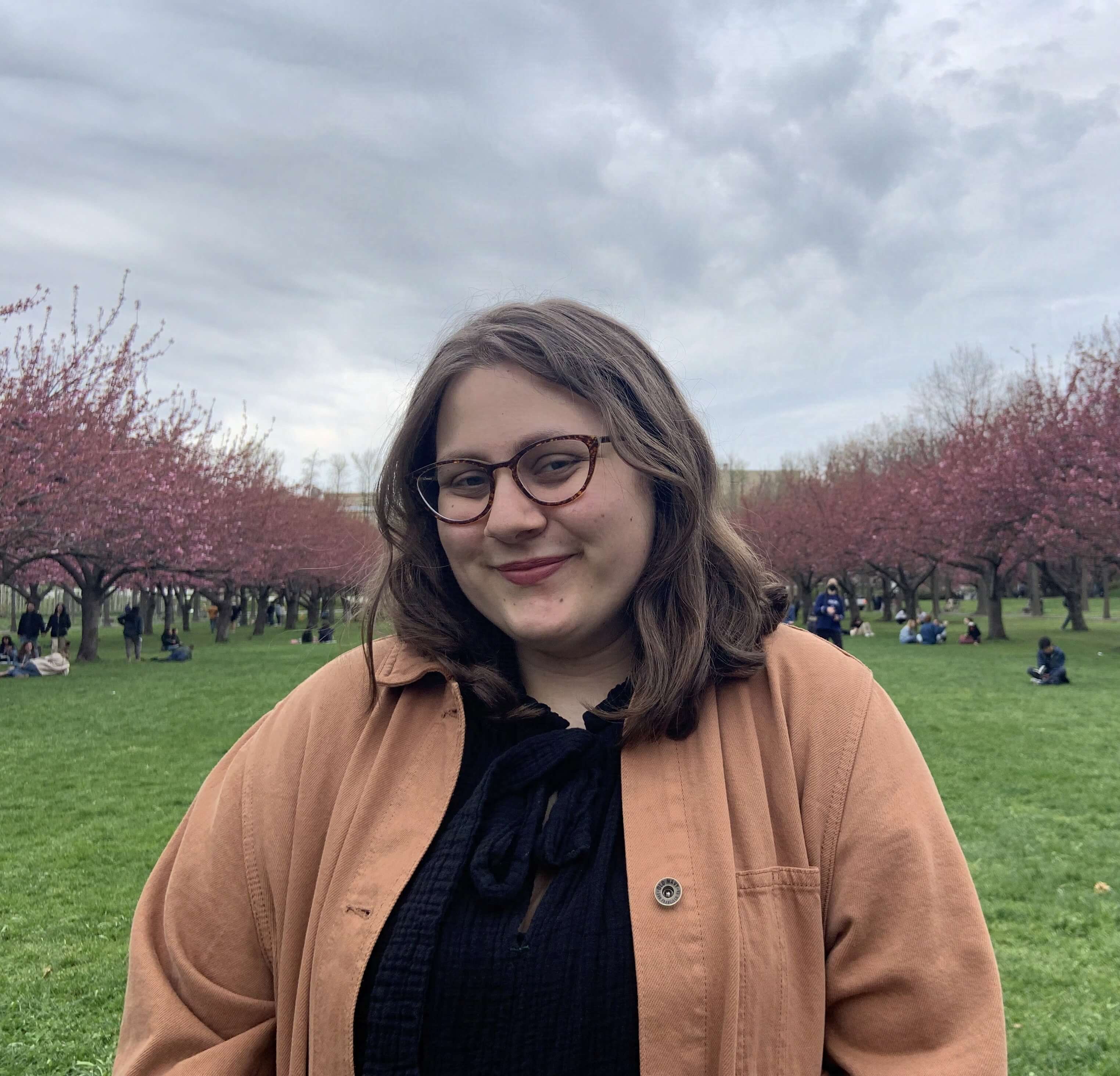 Headshot alt: Alin is a white person with brown hair and cateye glasses who is standing in a cherry blossom grove.