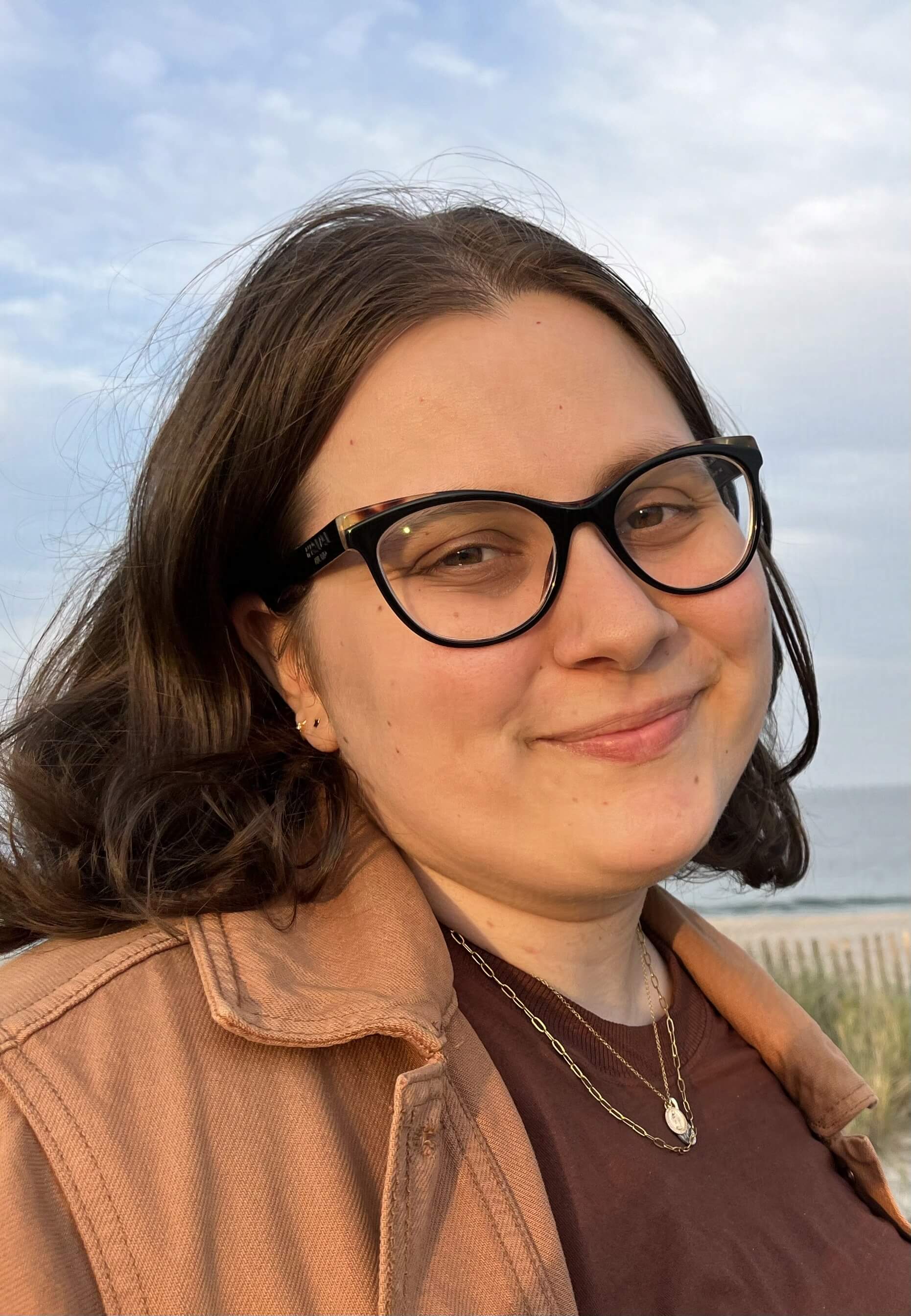 Headshot alt: Alin is a white person with brown hair and cateye glasses who is standing near a beach.